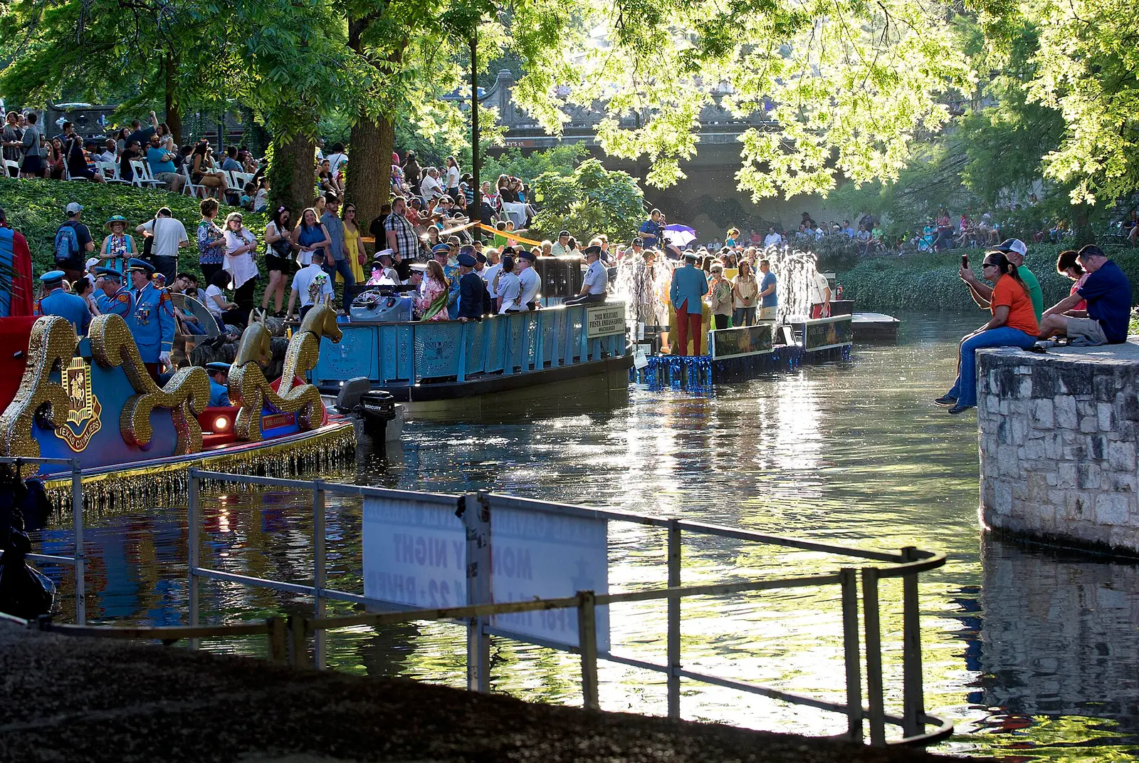 Decorated floats on the San Antonio River during Fiesta San Antonio, with crowds along the banks